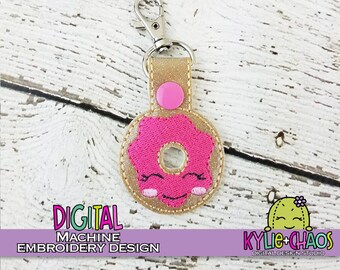 Può includere: Un portachiavi color oro con un design a forma di ciambella rosa. La ciambella ha una faccia sorridente ed è attaccata a un cinturino color oro con un bottone rosa. Il portachiavi ha un moschettone argentato. Il testo sull'immagine recita "DIGITAL MACHINE EMBROIDERY DESIGN".