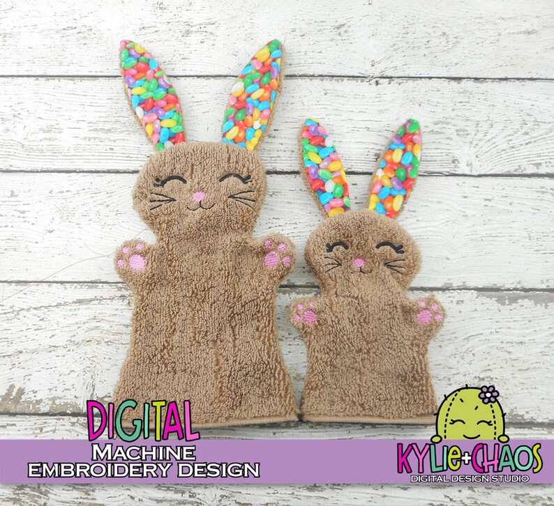 May include: Two brown bunny-shaped hand puppets with embroidered faces and pink paw pads. The bunny ears are filled with colorful jelly beans. The puppets are displayed on a white wooden surface. Text on the image reads "DIGITAL MACHINE EMBROIDERY DESIGN" and "KYLIE+CHAOS DIGITAL DESIGN STUDIO."