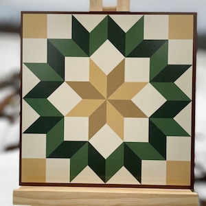 May include: A square, geometric wall art piece featuring a starburst design. The artwork uses shades of green, beige, and cream, with a dark brown border. The piece is displayed on a small wooden easel, perfect for home decor.
