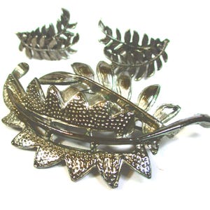 May include: A silver-toned brooch featuring a fern leaf design. The brooch is made of metal and has a textured finish. It comes with two smaller matching fern leaf earrings.