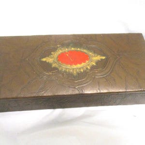 May include: A rectangular brown box with a textured surface. The lid features an embossed design with a central red oval framed by gold accents. The box appears to be made of a leather-like material.