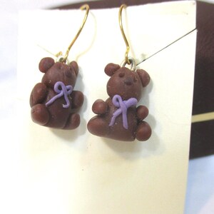May include: A pair of brown teddy bear earrings with purple bows. The earrings have gold-colored hooks. The bears are a dark brown color and have a cute, handmade appearance. The earrings are displayed on a white surface.