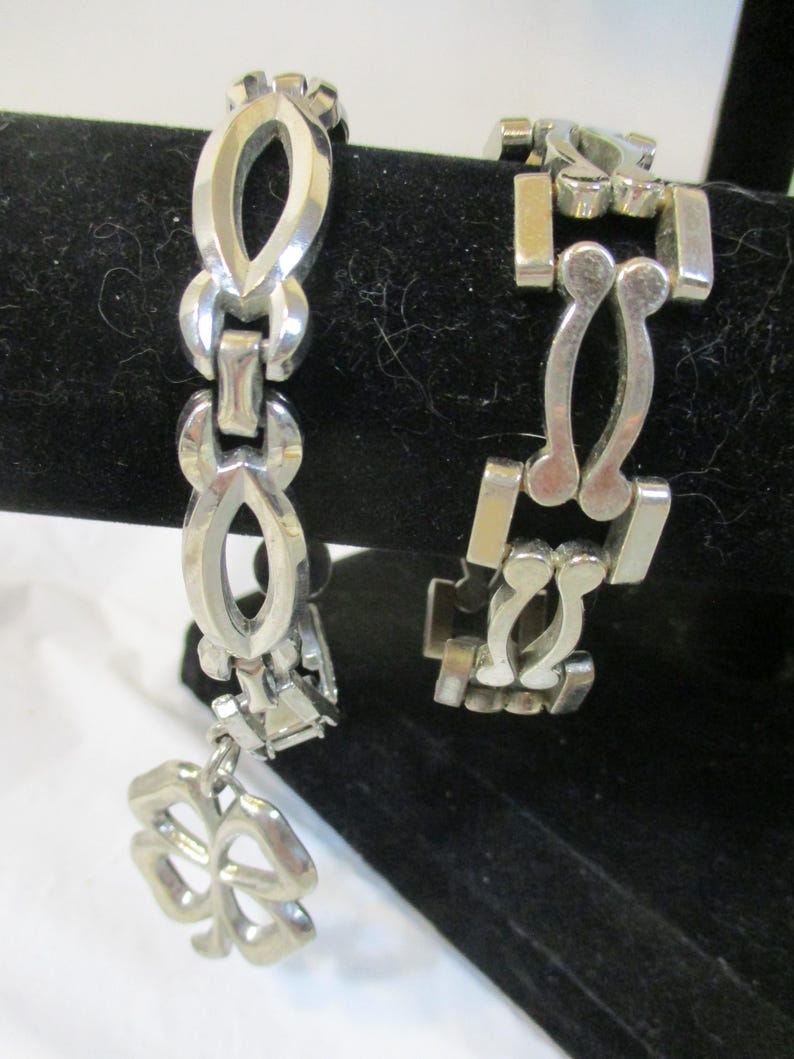 May include: Two silver bracelets. One has an open oval link design with a four-leaf clover charm. The other has a geometric design with rectangular and curved links. Both are displayed on a black surface.