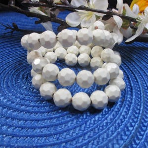 May include: A stack of three white beaded bracelets with faceted beads. The bracelets are on a blue textured surface, with white flowers and greenery in the background. The beads have a geometric design.
