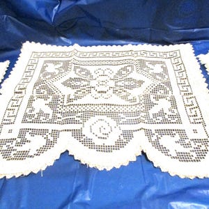 May include: Off-white, open-weave lace doilies with floral and geometric patterns. The doilies have a scalloped edge and are displayed on a blue surface. The lace has a delicate, handmade appearance.