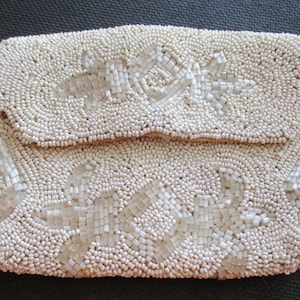 May include: A white beaded clutch bag with a floral design. The bag is made of small, round beads that are sewn together to create a textured surface.