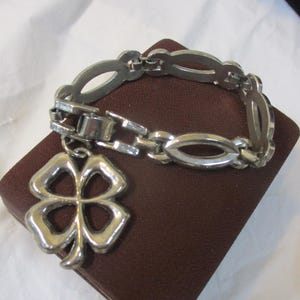 May include: Silver bracelet with a four-leaf clover pendant. The bracelet has an oval link design and a clasp. The clover charm is detailed and open. The bracelet is displayed on a brown, rectangular surface.