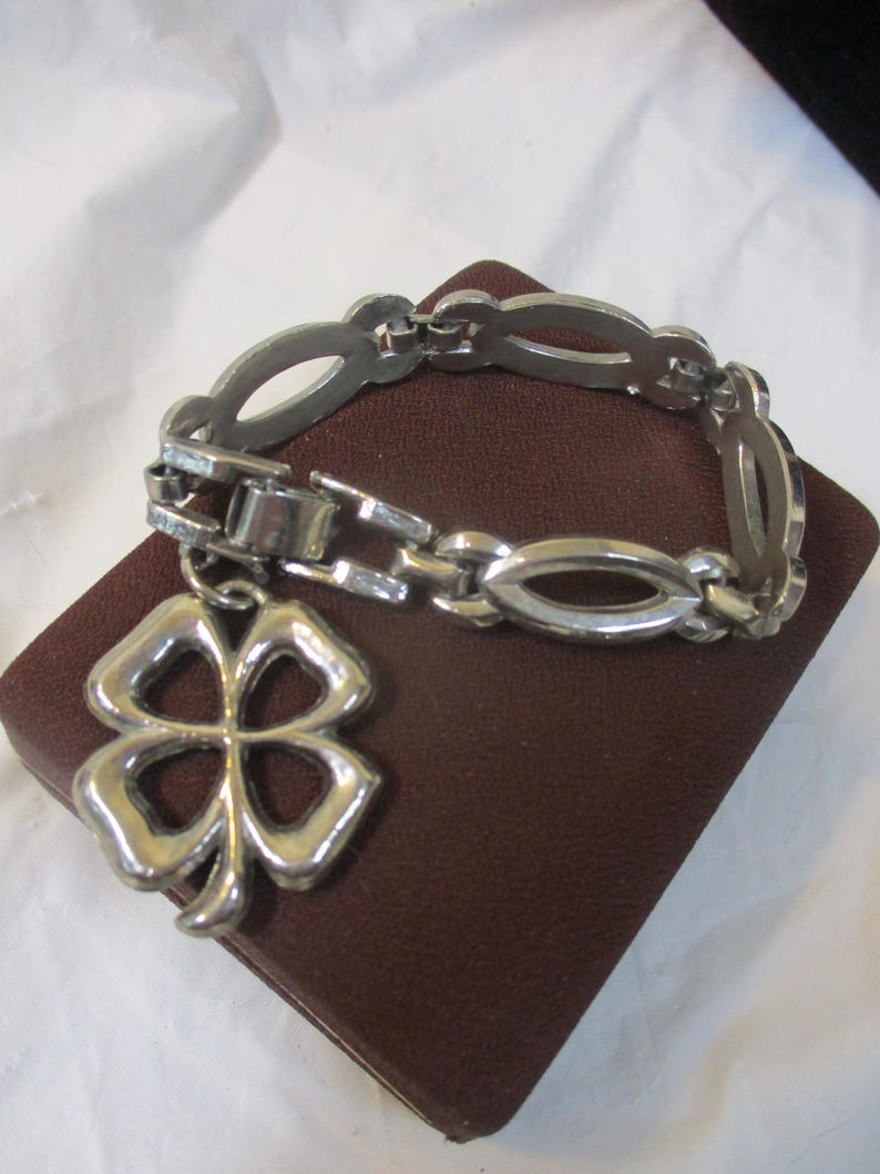 May include: Silver bracelet with a four-leaf clover pendant. The bracelet has an oval link design and a clasp. The clover charm is detailed and open. The bracelet is displayed on a brown, rectangular surface.