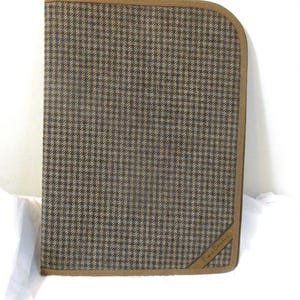 May include: A rectangular portfolio case featuring a brown, tan, and blue houndstooth pattern. The case has a rounded top right corner, a solid brown fabric trim along the edges, and a small brown triangular accent patch with the text Pierre Cardin.