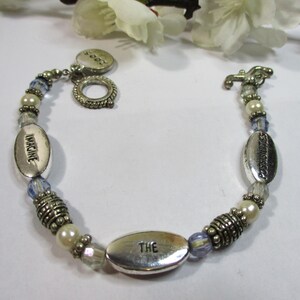 May include: A silver bracelet with blue and white beads, featuring oval charms with the words "Imagine", "The", and "Sunshine". The bracelet also has a heart charm with the word "Love" on it.
