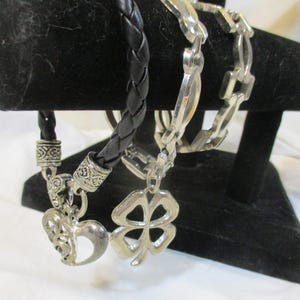 May include: Three bracelets on a black stand. One is a black, braided leather bracelet with a heart charm. Another is a silver chain bracelet with a clover charm. The third is a silver chain bracelet.