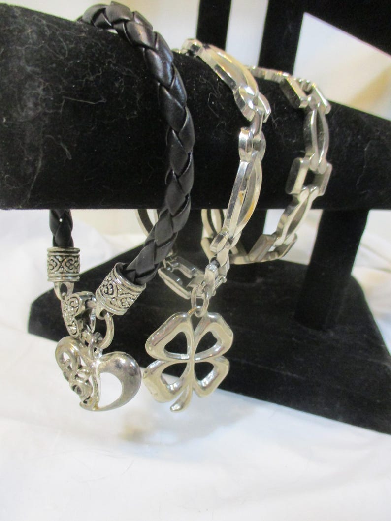 May include: Three bracelets on a black stand. One is a black, braided leather bracelet with a heart charm. Another is a silver chain bracelet with a clover charm. The third is a silver chain bracelet.