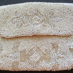 May include: A white beaded clutch bag with a floral design. The bag is made of small, round beads that are sewn together to create a textured surface. The floral design is created with larger, square beads that are arranged in a pattern.