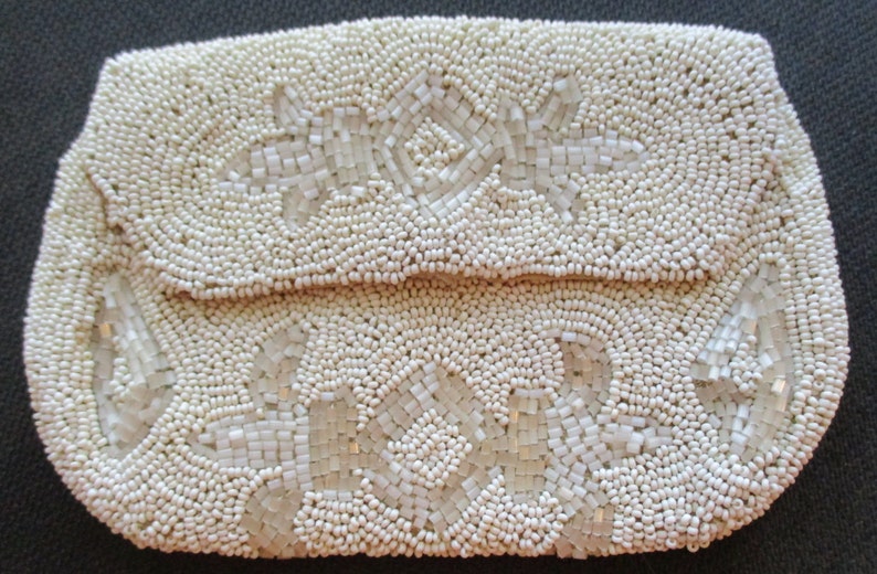 May include: A white beaded clutch bag with a floral design. The bag is made of small, round beads that are sewn together to create a textured surface. The floral design is created with larger, square beads that are arranged in a pattern.