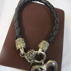 May include: Black braided leather bracelet with silver-coloured metal accents and a heart-shaped charm. The bracelet features a woven design and a decorative clasp. The heart charm has an open filigree design.