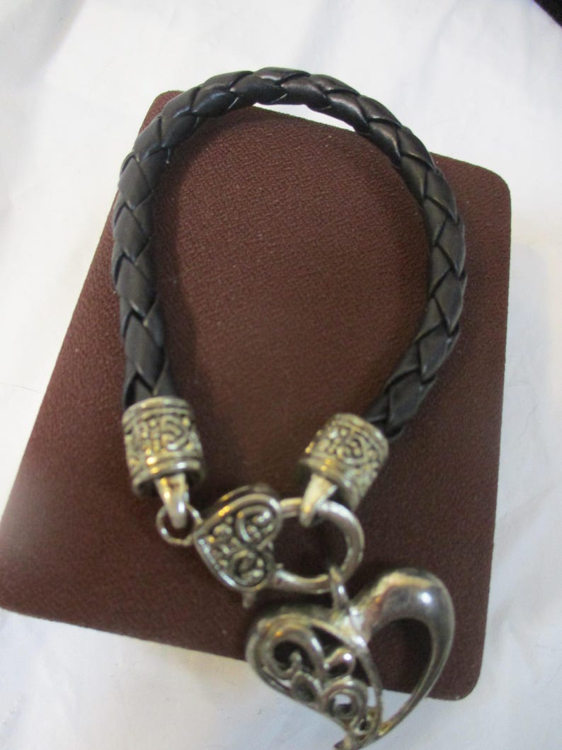May include: Black braided leather bracelet with silver-coloured metal accents and a heart-shaped charm. The bracelet features a woven design and a decorative clasp. The heart charm has an open filigree design.