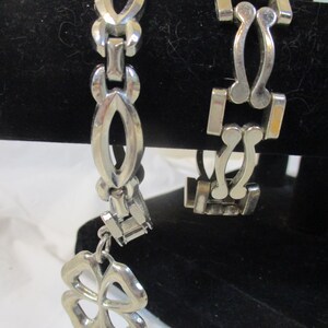 May include: Two silver bracelets. One has a clover charm. The other has a geometric design. Both are displayed against a black background. The bracelets are made of a shiny metal.