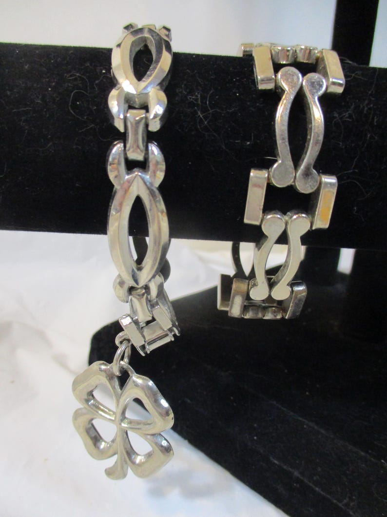 May include: Two silver bracelets. One has a clover charm. The other has a geometric design. Both are displayed against a black background. The bracelets are made of a shiny metal.
