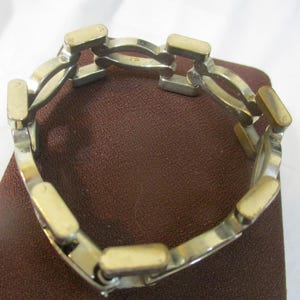 May include: Silver bracelet with gold-coloured rectangular accents. The bracelet features interconnected curved links and rectangular bars. The jewellery is displayed on a brown surface.
