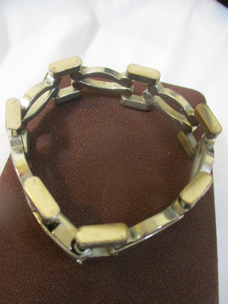 May include: Silver bracelet with gold-coloured rectangular accents. The bracelet features interconnected curved links and rectangular bars. The jewellery is displayed on a brown surface.