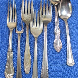 May include: A set of vintage silverware, including forks and spoons, with ornate handles. The silverware is silver-plated and has a polished finish. The silverware is arranged on a blue background.