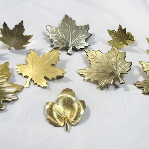 May include: A collection of decorative leaf-shaped objects in various metallic finishes, including gold and silver. The leaves are detailed with veining and have a three-dimensional appearance. The arrangement suggests a fall or nature-inspired theme.