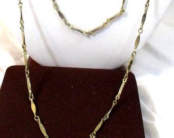 May include: Two gold-toned necklaces. The longer necklace is draped, while the shorter one rests above it. Both feature a repeating pattern of elongated, oval-shaped links connected by smaller, circular links. The necklaces are displayed on a dark maroon surface.