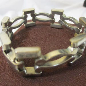 May include: A silver-coloured bracelet with a distinctive link design. The bracelet features rectangular and curved links, creating an open, geometric pattern. The jewellery is displayed on a brown surface.