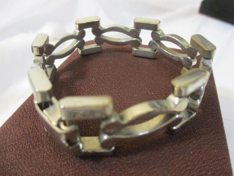 May include: A silver-coloured bracelet with a distinctive link design. The bracelet features rectangular and curved links, creating an open, geometric pattern. The jewellery is displayed on a brown surface.