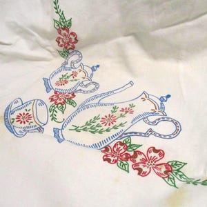 May include: White fabric with embroidered floral and teapot design. The embroidery features red and green flowers, blue accents, and a teapot and cup motif. The design is likely for a tablecloth or similar textile.