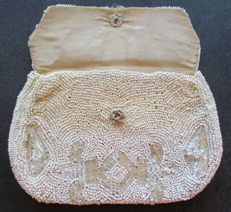 May include: A white beaded clutch bag with a floral design. The bag is open and the inside is lined with a cream-coloured fabric.