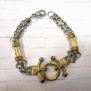 May include: A silver and gold-tone bracelet with a unique design. The bracelet features a double chain, gold-tone cylindrical accents, and a central clasp with a circular element and decorative arms. Small blue and red accents are visible.
