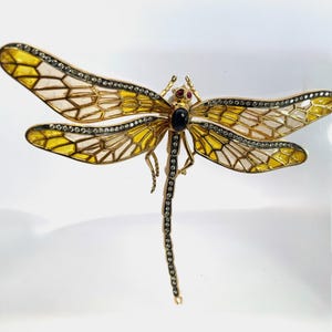 May include: A gold-toned dragonfly brooch featuring translucent yellow and white enamel wings outlined with small clear crystals. The body is accented with a dark oval stone at the center, two small red stones for eyes, and a long tail set with crystals.