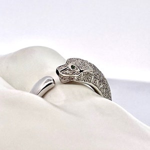 May include: A silver ring with a diamond-encrusted snake design. The snake's head is facing the viewer, and its eyes are green gemstones. The ring is on a white surface.