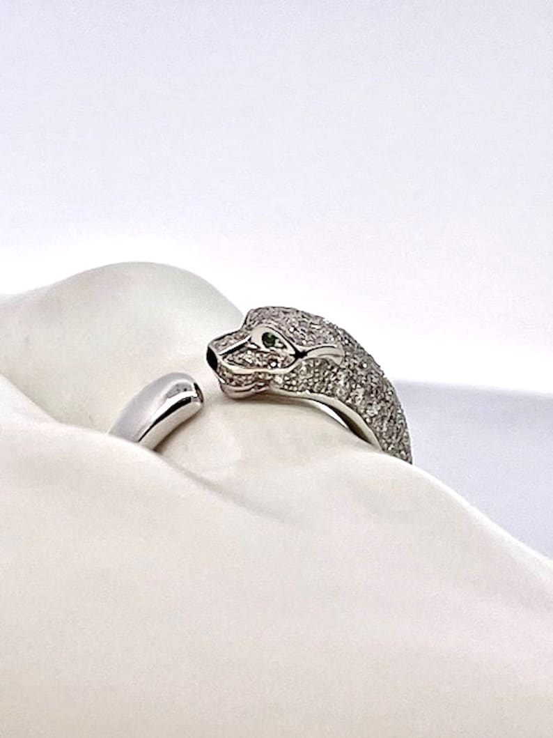 May include: A silver ring with a diamond-encrusted snake design. The snake's head is facing the viewer, and its eyes are green gemstones. The ring is on a white surface.