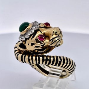 May include: A gold ring with a tiger head design. The tiger has black stripes, red ruby eyes, and a green emerald on its head. The ring is set with diamonds around the emerald.