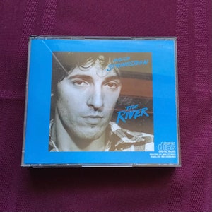 May include: A blue CD case with a photo of Bruce Springsteen on the front. The title of the album is "The River" and the text "Compact Disc Digital Audio Digitally Mastered Analog Recording" is printed on the bottom right corner.