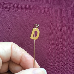 May include: A gold-coloured letter D charm on a thin gold-coloured stick pin.