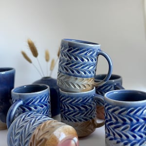 May include: A collection of handmade ceramic mugs in shades of blue and tan. The mugs feature a textured leaf pattern and curved handles. Some mugs are stacked, showcasing the variety of colors and designs. A vase and dried plants are in the background.
