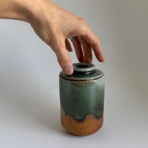 以下が含まれることがあります： 蓋付きの陶器の壺。壺は円筒形で、緑色の釉薬が施され、茶色のベースにフェードします。蓋は濃い緑色で、黒い縁があります。壺は手で持たれています。