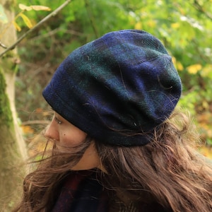 以下が含まれることがあります： 青、緑、黒のチェック柄ビーニー帽と、お揃いのスカーフ。 スカーフはチェック柄のシャツとグリーンのコートの上にかけられています。 背景は、緑と黄色の葉のある森のぼやけた景色です。