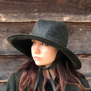 May include: A woman wearing a dark green coat with double breasted buttons and a wide brimmed green hat with a black ribbon tied under her chin.