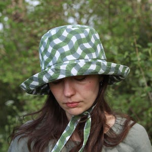 May include: A woman wearing a green and white checked bucket hat with a matching green and white checked ribbon tied under her chin. The hat has a wide brim and is made of a soft, lightweight fabric.