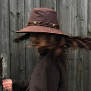 Puede incluir: Una persona lleva un sombrero de fieltro marr&oacute;n con un ala ancha y detalles decorativos. El sombrero tiene una banda marr&oacute;n oscura con detalles dorados. La persona lleva un jersey de cuello alto marr&oacute;n y tiene el pelo largo y oscuro.