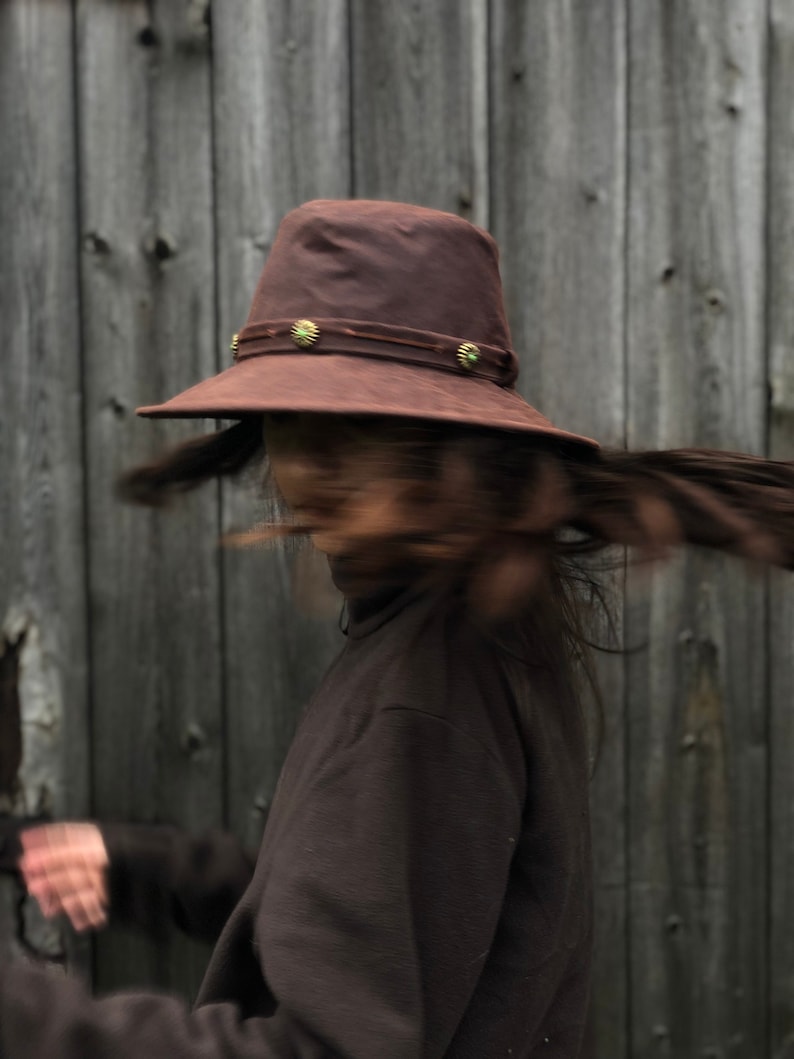 Puede incluir: Una persona lleva un sombrero de fieltro marr&oacute;n con un ala ancha y detalles decorativos. El sombrero tiene una banda marr&oacute;n oscura con detalles dorados. La persona lleva un jersey de cuello alto marr&oacute;n y tiene el pelo largo y oscuro.