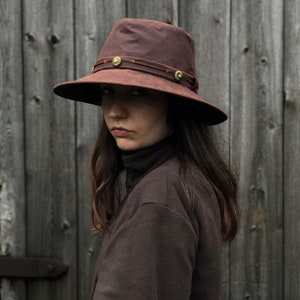 Puede incluir: Un sombrero de lona encerada marr&oacute;n con un ala ancha y una banda de cuero con detalles met&aacute;licos decorativos. El sombrero lo lleva una persona con el pelo largo y casta&ntilde;o.