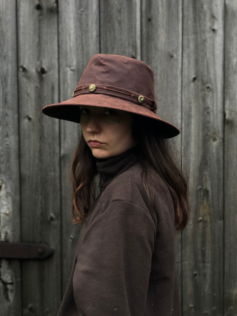 Puede incluir: Un sombrero de lona encerada marr&oacute;n con un ala ancha y una banda de cuero con detalles met&aacute;licos decorativos. El sombrero lo lleva una persona con el pelo largo y casta&ntilde;o.
