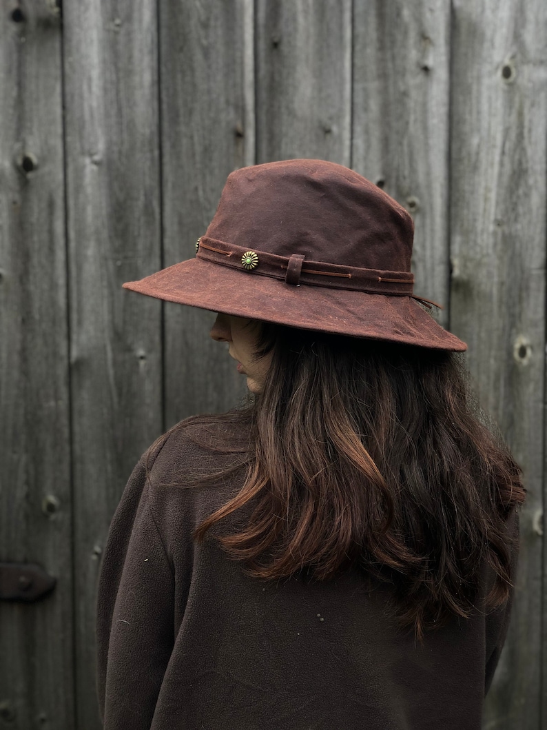 Puede incluir: Un sombrero marr&oacute;n de ala ancha con una banda de cuero y un peque&ntilde;o acento de flor de metal. El sombrero lo lleva una persona con el pelo largo y casta&ntilde;o.