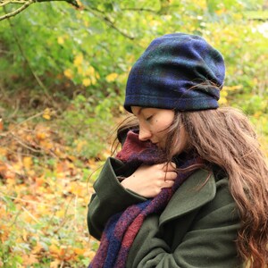 May include: A dark green coat, a blue and green plaid beanie, and a scarf with blue and burgundy plaid patterns. The scarf is wrapped around the neck. The background is a blurred view of a forest with autumn leaves.