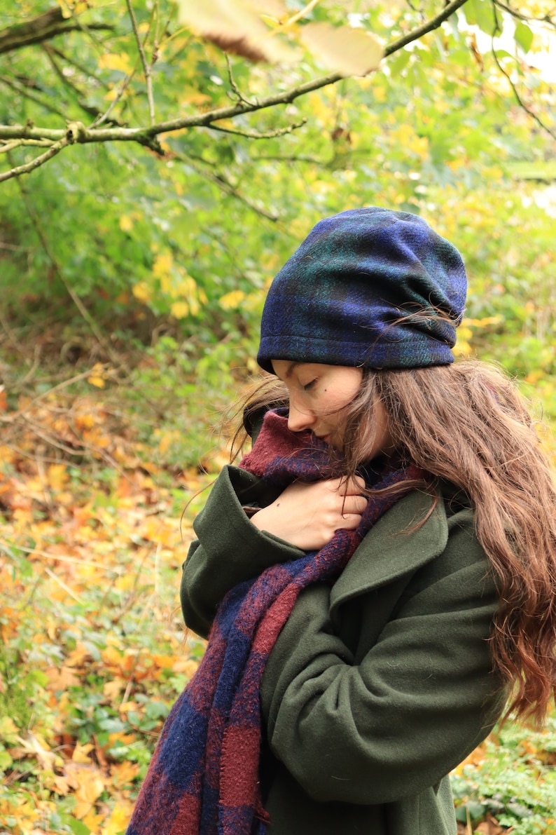 May include: A dark green coat, a blue and green plaid beanie, and a scarf with blue and burgundy plaid patterns. The scarf is wrapped around the neck. The background is a blurred view of a forest with autumn leaves.